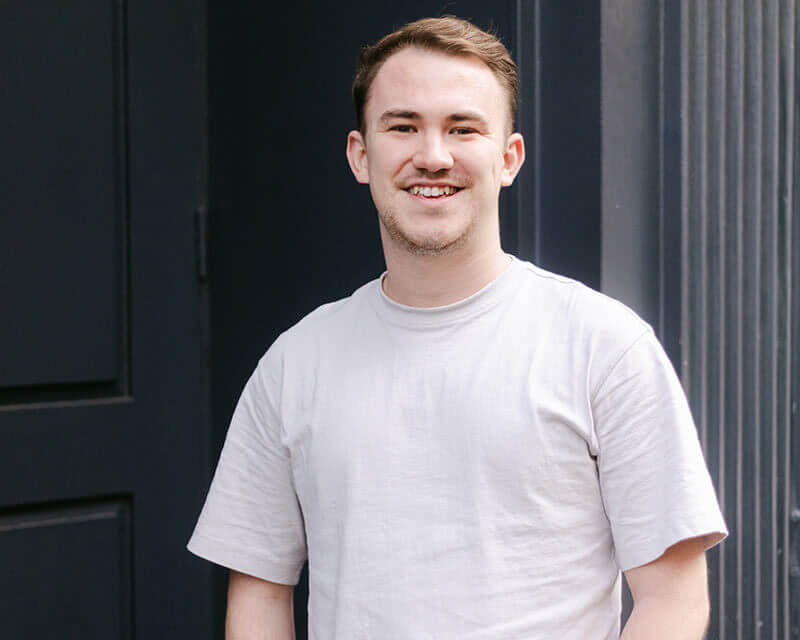 Man wearing a light beige t-shirt, smiling and standing in front of a Black doorway