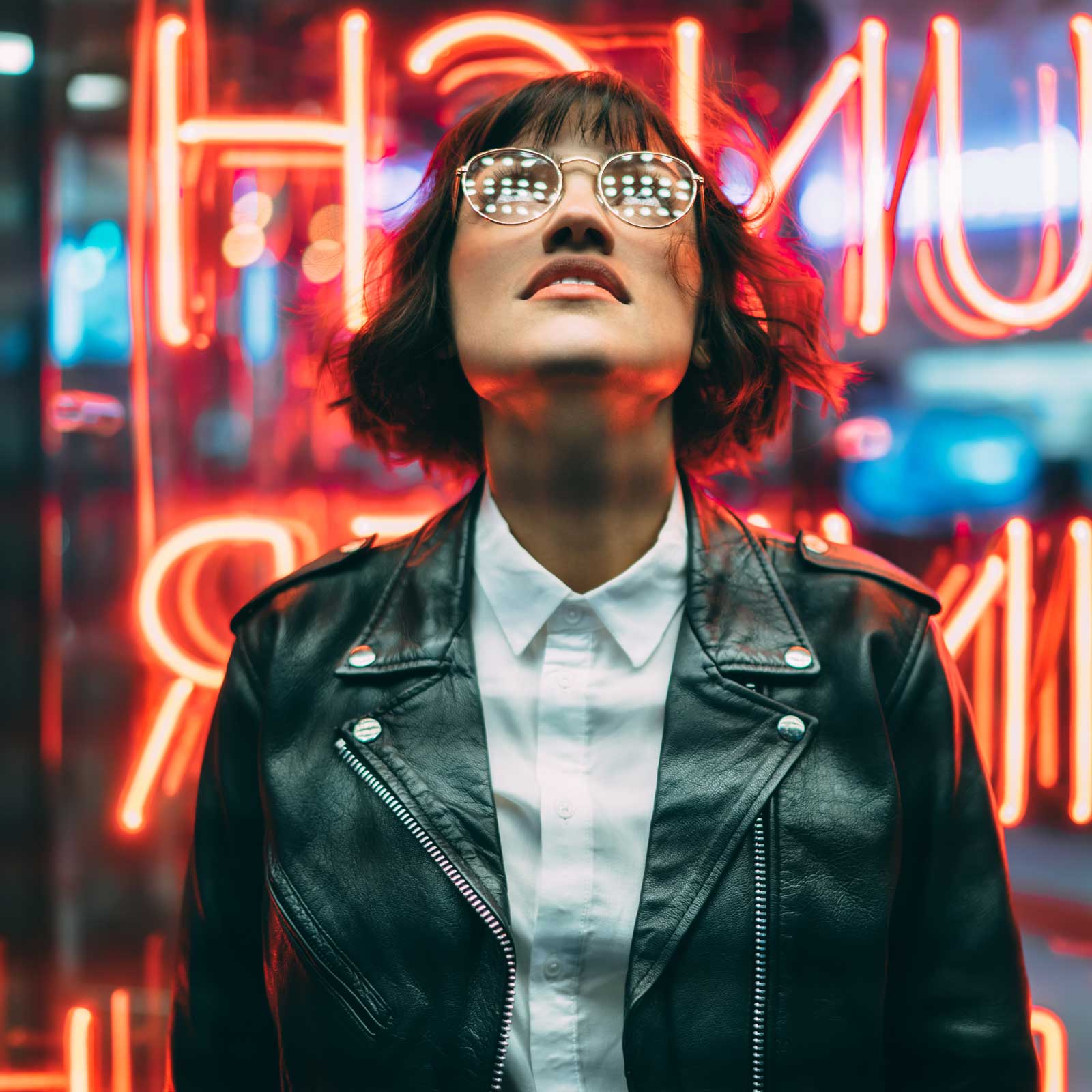 Person wearing glasses and a leather jacket looking up and standing in front of red neon lights