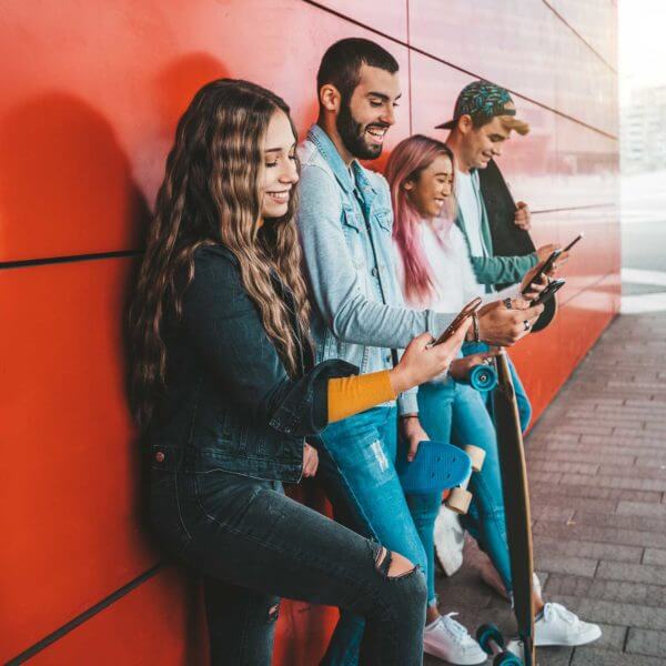 4 young people all looking at their smartphones leaning up against a red wall