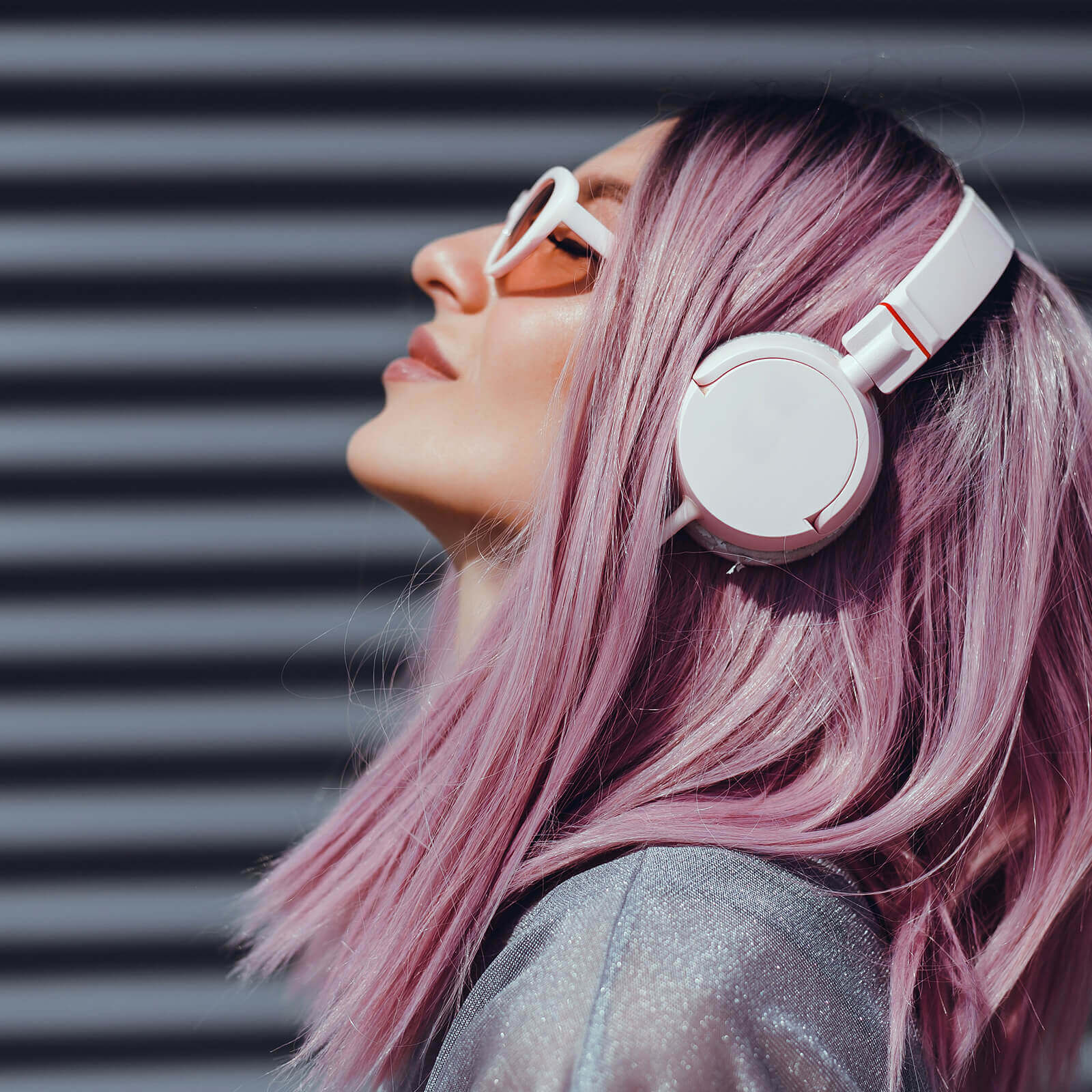 Woman with lilac hair and sunglasses tipping head back and listening to headphones.
