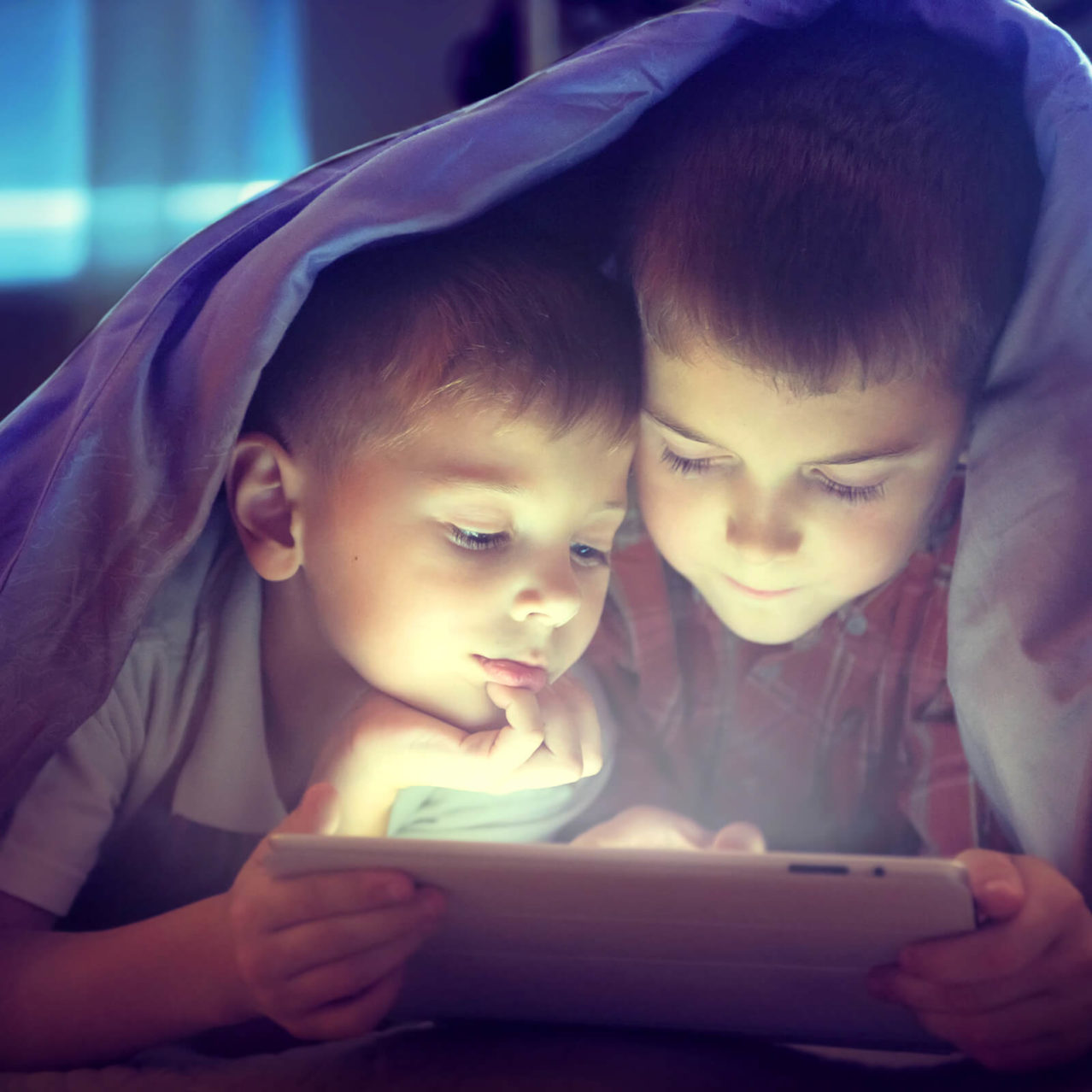 2 kids watching a tablet under a duvet