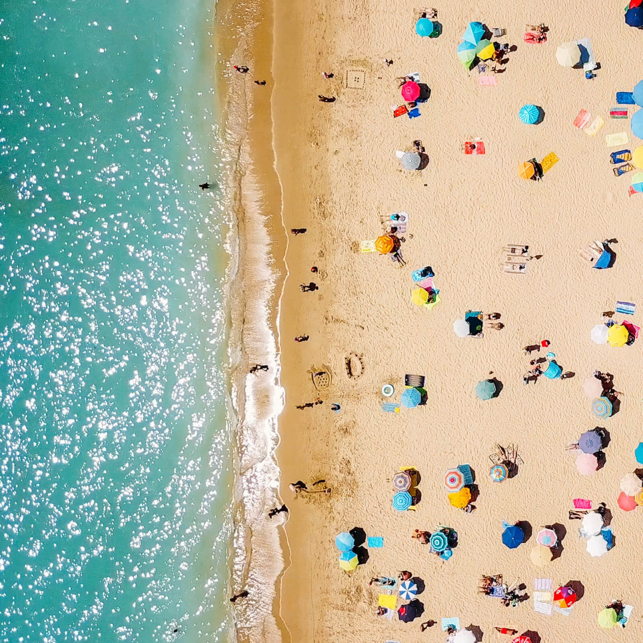 Birds eye view of a busy beach resort, colour umbrellas, golden sand and turquoise water.