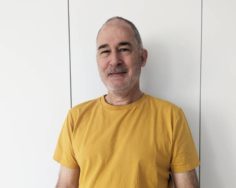 Man wearing a yellow t-shirt stood against a white wall.
